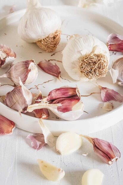 Fresh garlic cloves and bulbs arranged on a plate with scattered garlic skin on a white surface