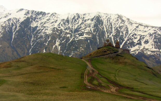 Snow capped peaks behind green hills in Georgia landscape with old structures on top