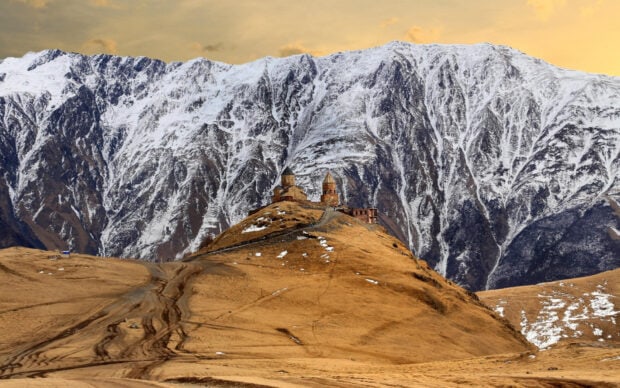 Ancient Georgia church on brown hill with snow covered mountains in the background