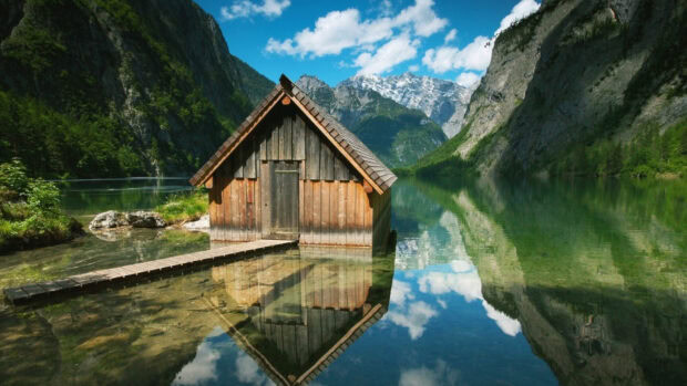 A wooden cabin surrounded by mountains and clear water in Germany reflecting the blue sky and clouds