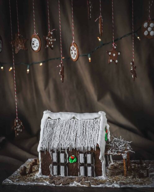 A detailed gingerbread house with iced roof and hanging decorated cookies in the background