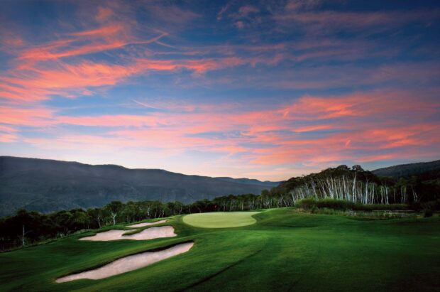 A golf course surrounded by trees and hills under a colorful sunset sky