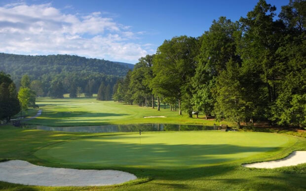 A golf course surrounded by trees and hills with a sand bunker and a flag on the green