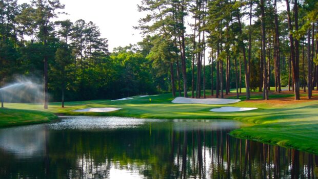 A golf course surrounded by trees with a water hazard and sand bunkers on the green