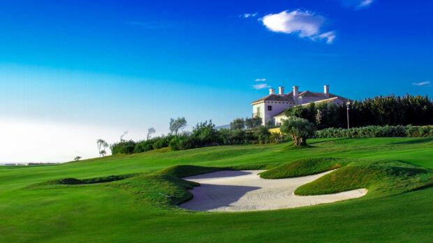 A golf course with green grass and a sand trap near a large house under a clear blue sky