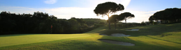 A golf course with sand bunkers and trees under a clear sky in the late afternoon sunlight