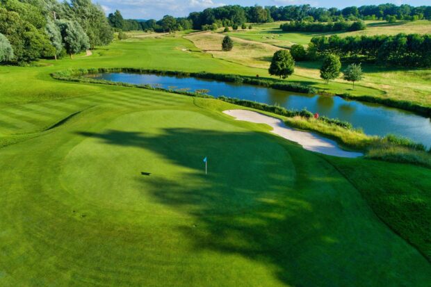 A golf course with a green and water hazard under a clear blue sky with surrounding trees