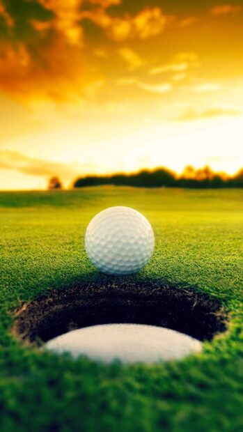 A golf ball is positioned on the edge of a hole on a golf course at sunset