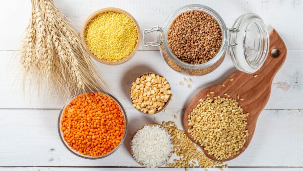 Various grains displayed in bowls and jars on a white wooden surface with ears of wheat