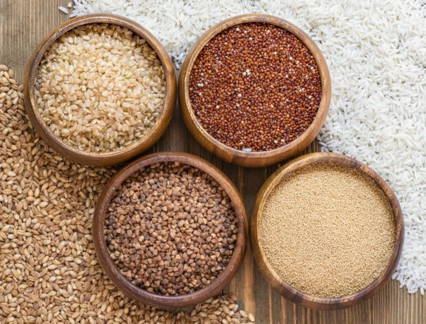 Various grains arranged in wooden bowls on a wooden surface including quinoa buckwheat and rice