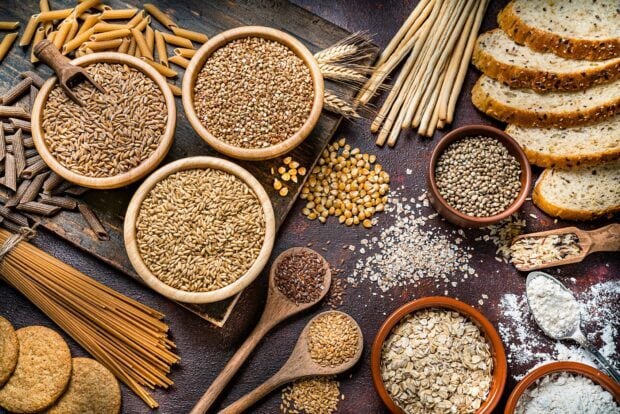 Various grains in wooden bowls and spoons on a rustic table with bread and pasta