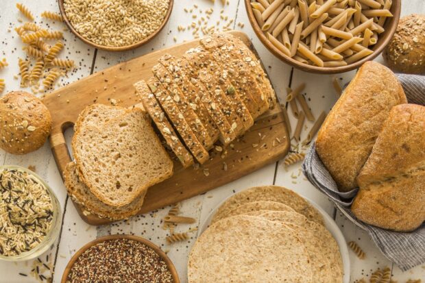 A variety of grains displayed with sliced multigrain bread and whole grain pasta on a wooden surface