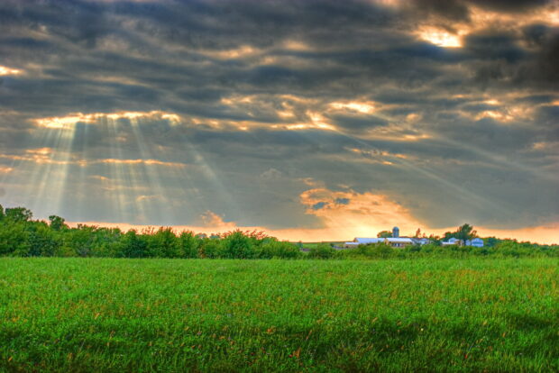 Sun rays shining through clouds over a vast grassland with a farmhouse in the distance