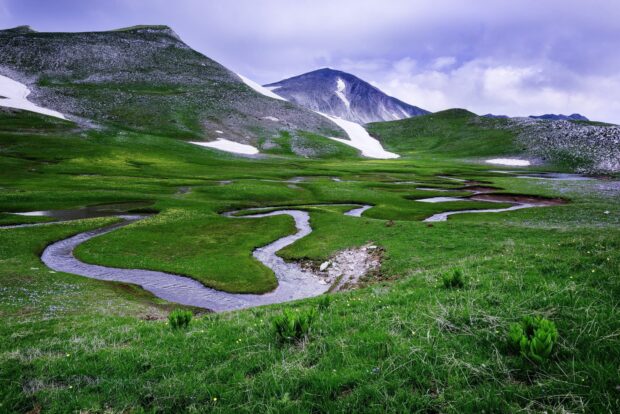 A green grassland with winding streams flowing through mountainous terrain and patches of snow