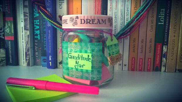 A gratitude jar decorated with colorful beads placed on a desk with a pink pen and books in the background