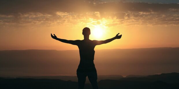 A person expressing gratitude with arms outstretched at sunset over mountains