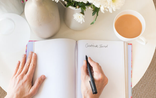 Person writing in gratitude journal on white table with coffee and flowers