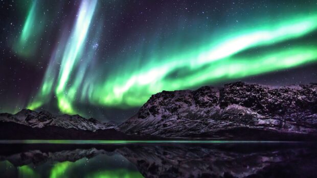 Green northern lights over snowy mountains in Greenland reflecting on calm water