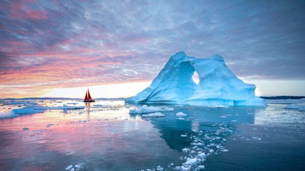 A sailboat near a large iceberg with a natural arch in Greenland at sunset