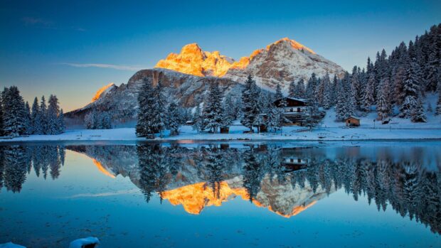 Snowy Greenland mountains illuminated by sunset reflected on calm lake with pine trees
