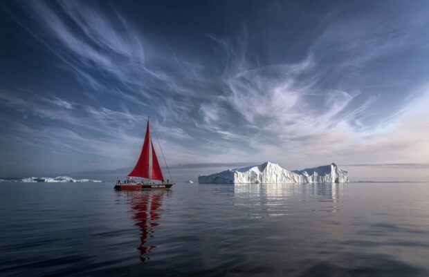 A sailboat with red sails near a large iceberg in Greenland calm waters