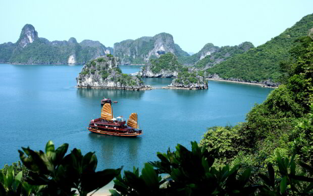 Traditional wooden boat sailing near limestone islands in Hanoi bay with calm blue water and green trees