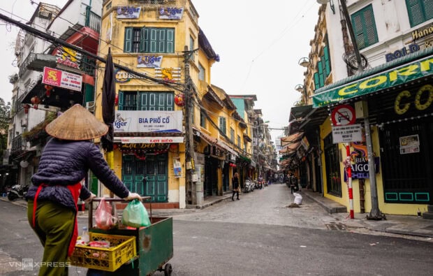 A street vendor wearing a conical hat carries goods on a cart in the Old Quarter of Hanoi