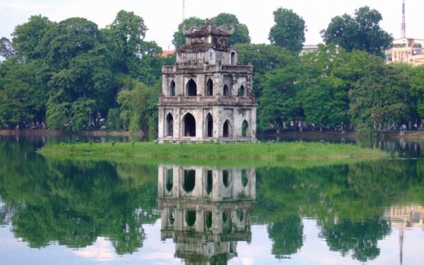 Ancient tower in Hanoi surrounded by lush greenery and calm lake reflecting the structure