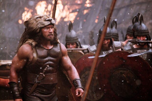 Hercules wearing a lion head and armor walking in a battle scene with soldiers behind him