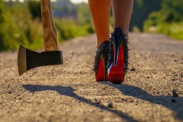 Woman wearing high heels walking on a dirt road next to an axe standing upright