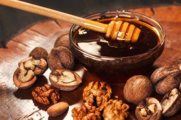 A bowl of honey with a wooden dipper surrounded by cracked and whole walnuts on a wooden surface