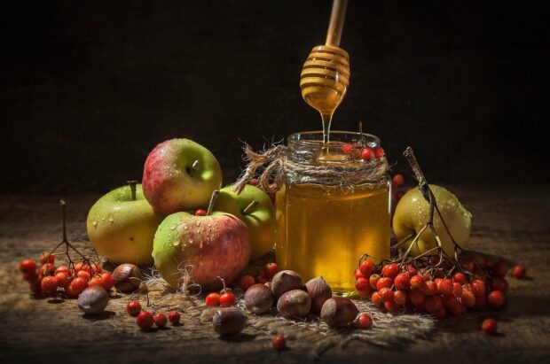 A jar of honey with apples and red berries on a rustic wooden surface