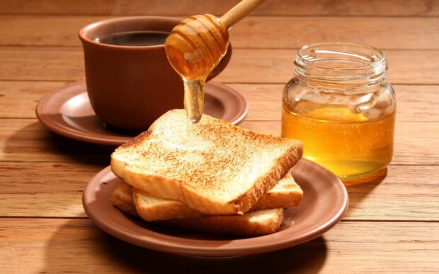 Honey being drizzled on toasted bread slices with a jar of honey on wooden table