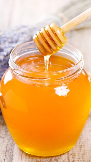 A wooden honey dipper dripping golden honey into a glass jar of honey