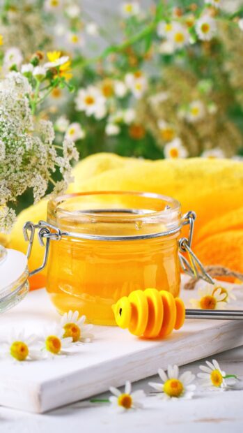 A jar of honey with chamomile flowers and a honey dipper on a white wooden board