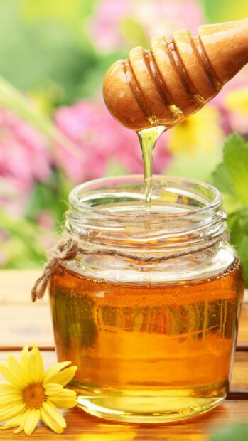 Golden honey dripping from wooden dipper into glass jar of honey