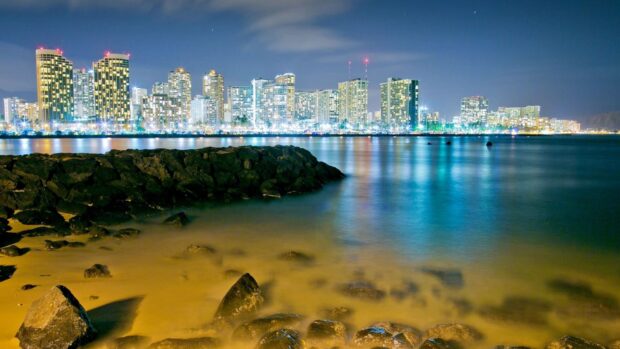 Night view of Honolulu city skyline with illuminated buildings and rocky shore