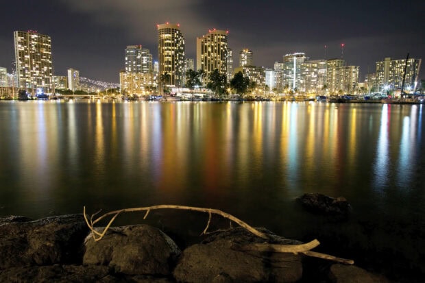 Nighttime cityscape of Honolulu with illuminated buildings reflecting over calm water