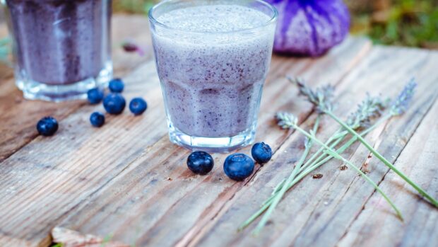 A fresh huckleberry smoothie glass on a wooden table with scattered huckleberries and purple flowers