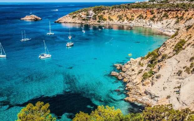 Clear turquoise waters and rocky cliffs in Ibiza bay with anchored sailboats
