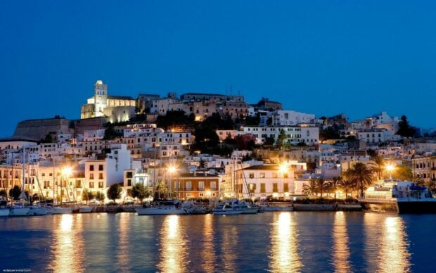 Illuminated historic Ibiza town by the sea at night with boats docked in the harbor