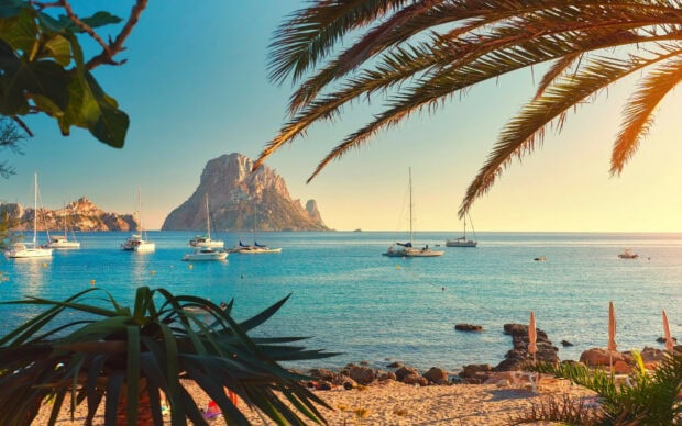 A scenic view of Ibiza coastline with sailboats and rocky island under clear blue sky