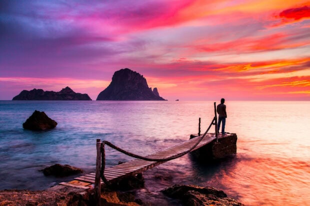 A person standing on a wooden pier at sunset with Ibiza rock formations in the distance