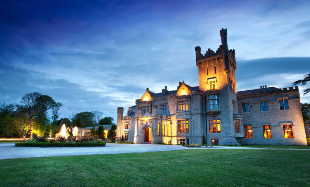 An illuminated Irish castle at dusk surrounded by green lawn and blue sky