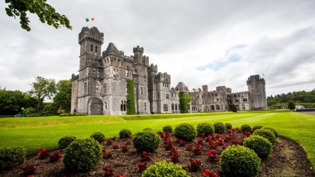 Irish castle surrounded by green bushes and red flowers under a cloudy sky