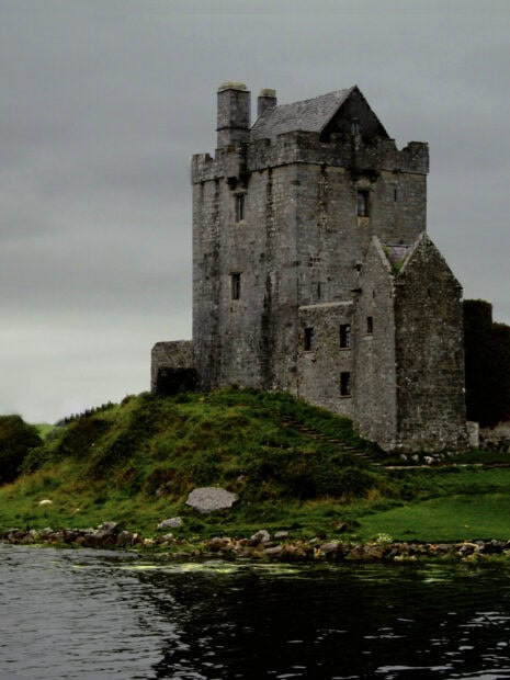 Ancient Irish castle sits on a grassy hill near water under cloudy skies