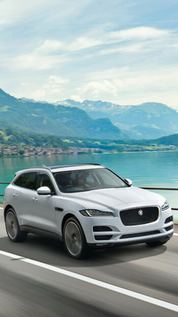 A white Jaguar car driving on a road with mountains and lake in the background