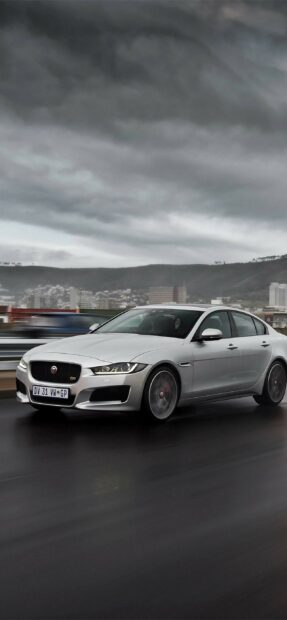 A silver Jaguar car driving on a wet road under a cloudy sky in an urban area