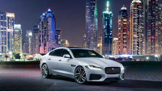 A silver Jaguar car parked in front of a city skyline at night featuring tall illuminated towers