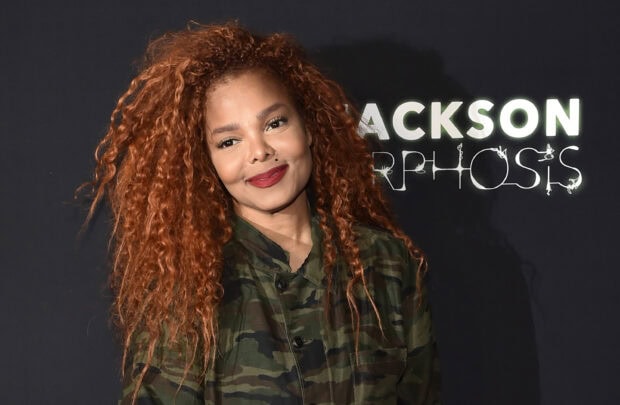 Janet Jackson wearing camouflage jacket with long curly hair and red lipstick smiling